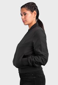 Schwarze Bomberjacke mit Reißverschluss, gerippten Bündchen und einem Aufnäher auf der Brust. Sie verfügt über einen glatten, leichten Stoff und eine strukturierte Passform.