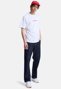 Hvid bomulds-t-shirt med rødt logo, parret med mørkeblå denimjeans og hvide sko. Rød kasket tilføjer en koordinerende farveaccent.