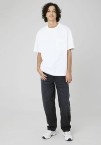 T-shirt en coton blanc avec une poche, associé à un pantalon en denim foncé. Le modèle porte également des chaussures de sport blanches. Fond uni.