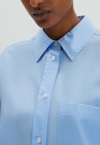 Camicia azzurro chiaro con bottoni, realizzata in tessuto morbido e con colletto a punta, dotata di bottoni in madreperla e una tasca frontale.