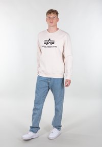 Felpa rosa chiaro in misto cotone con logo nero, abbinata a jeans azzurri e sneakers bianche. Collo rotondo semplice, vestibilità comoda.