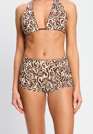 Frau trägt einen braun-cremefarbenen Neckholder-Bikini mit abstraktem Muster und passende hoch taillierte Badeshorts vor einem einfarbigen Hintergrund.