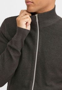 Sweatshirt zippé noir avec un col haut, des poignets côtelés et un tissu en maille texturée. Il présente une fermeture éclair contrastante de couleur gris clair.