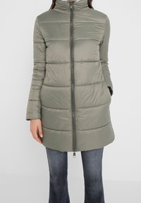 Kvinna klädd i knälång olivgrön quiltad dunjacka med blixtlås framtill och händerna i fickorna, kombinerad med blekta blå jeans.