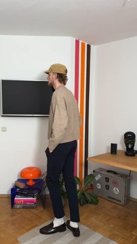 Bézs kötött pulóver, tengerészkék nadrág, barna bőrcipő és fehér zokni. A háttérben TV, narancssárga lámpa és színes csíkok a falon.