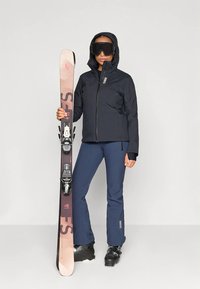 Frau in schwarzer Skijacke, blauen Skihosen und Stiefeln, die einen rosa und schwarzen Ski aufrecht hält. Sie trägt schwarze Skibrille und Kapuze.