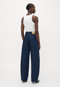 Jeans in denim scuro a gamba larga con vita alta, dotati di tasche posteriori e una patch in pelle con logo sulla cintura. Abbinati a un top bianco senza maniche.