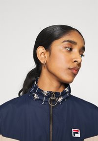 Giacca a vento blu marina con colletto decorato, chiusura con zip e patch con logo. Presenta un design semplice e una texture liscia.