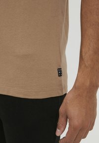 Brun bomull t-skjorte med en sideetikett som har "DNWORKR," kombinert med svarte bukser. Stoffet har en myk tekstur og en avslappet passform.