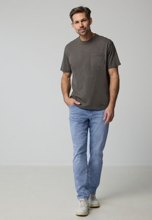 Man die vooruit loopt met een donkergrijs t-shirt met zak, lichtblauwe spijkerbroek en witte sneakers tegen een effen grijze achtergrond.