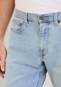 Los jeans de mezclilla azul claro presentan una cintura alta, un diseño clásico de cinco bolsillos, sutiles desgastes y costuras en contraste con un cierre de botón.