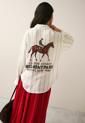 Femme aux longs cheveux foncés portant une chemise blanche rayée avec un motif cheval et jockey ainsi qu'un texte rouge, associée à une longue jupe rouge.
