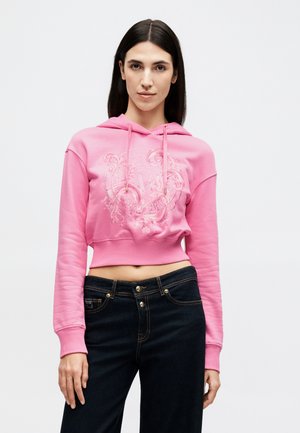 Žena s rovnými tmavými vlasy, která má na sobě krátkou růžovou mikinu s reliéfním vzorem a tmavé džíny s vysokým pasem, stojící před bílým pozadím.