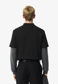 Polo negro de manga corta superpuesto sobre una camisa de manga larga gris. Pantalones negros de corte Slim con un cinturón. Textura de tela sutilmente visible.