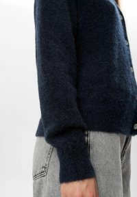 Mornarsko modra puhasta jopica z gumbastim zapiranjem, rebrastimi manšeti in mehko teksturo, nosena čez svetlo sivo jeans.