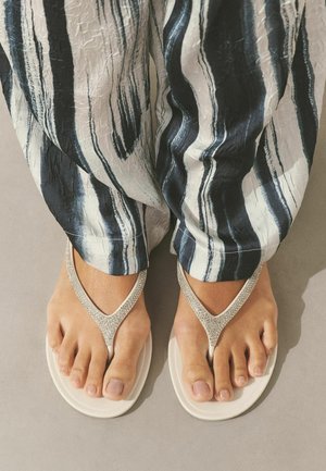 Lábak fehér, strasszos pántú flip-flop papucsban, laza, fekete-fehér csíkos nadrággal párosítva, sima szürke felületen állva.