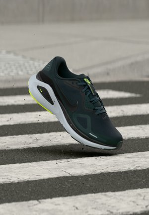 Chaussure de course Nike vert foncé et noire avec semelle blanche et jaune néon, prise en plein air au-dessus d'un passage piéton sur l'asphalte.