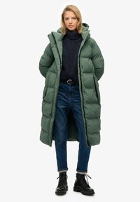 Langer grüner Daunenmantel mit hohem Kragen, vorderer Knopfleiste, großer Kapuze und Seitentaschen, über einem dunklen strukturierten Pullover und blauen Jeans kombiniert.