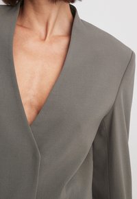 Blazer grigio con scollatura a V profonda, spalle strutturate e texture liscia, che mette in evidenza cuciture pulite e un design minimalista.