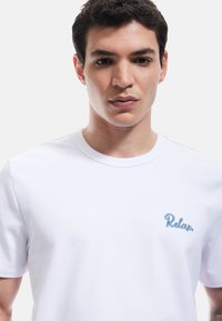Hvid bomulds t-shirt med runder hals og korte ærmer, udsmykket med lyseblå broderi med teksten "Relax." på venstre bryst.