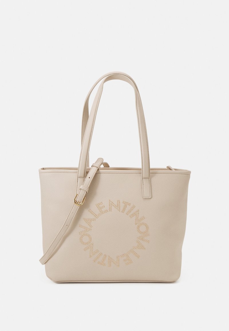 Beige läderväska med dubbla handtag, justerbar axelrem och cirkulär logotypdetalj prydd med nitar på framsidan.