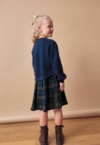 Marinblå sweatshirt, rutmönstrad kjol i mörkgröna och blå nyanser, robusta bruna stövlar, texturerat hår, stående i profil mot en beige bakgrund.