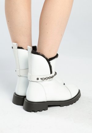 Valkoiset puolivartiset saappaat, joissa musta turvalaite ja ketjuyksityiskohta, henkilö käyttää ja seisoo valkoisella pinnalla.