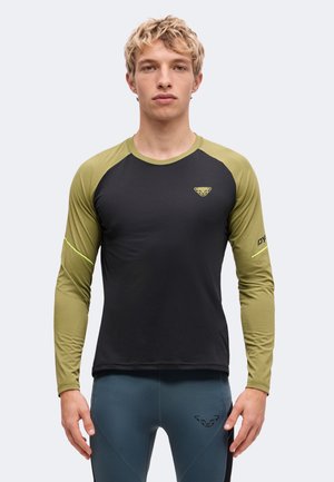 Junger Mann mit kurzen blonden Haaren, der ein schwarzes und olivgrünes langärmeliges Sportshirt sowie blaue enge Leggings trägt und nach vorne schaut.