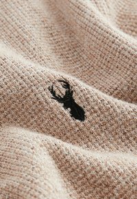 Beige strikket stof med et tekstureret mønster og en sort broderet hjortehorn accent. Materialet fremstår blødt og varmt.