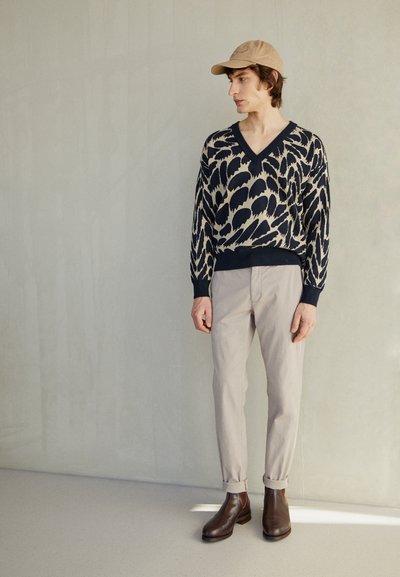 Granatowy sweter z kremowym wzorem liści, dekolt w serek, w zestawieniu z jasnobeżowymi spodniami i ciemnobrązowymi skórzanymi botkami do kostek. Dodatki: beżowa czapka.