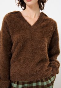 Brauner, flauschiger Pullover mit V-Ausschnitt, weichem Material und lockerer Passform, getragen mit grün-braun karierter Hose.