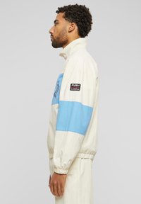 Cream and light blue track jacket with a zipper, featuring a blue stripe, elastic cuffs, FUBU logo patch on the arm, and lightweight fabric.