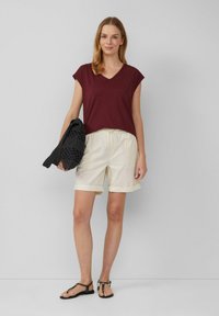Bordowy t-shirt z dekoltem w serek i krótkimi rękawami, połączony z beżowymi szortami z podwiniętymi nogawkami; trzyma czarną dzianinową torbę; nosi czarne sandały.