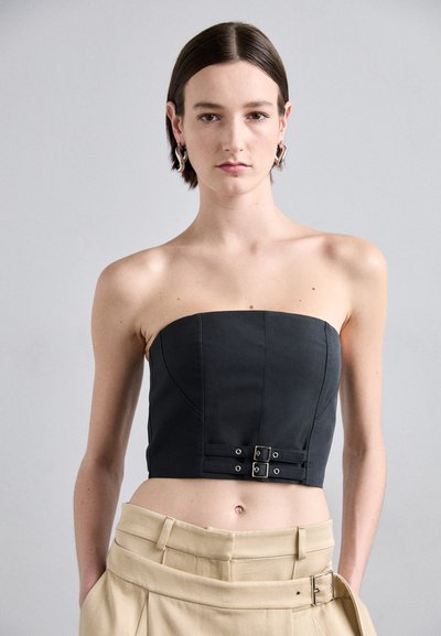 Černý bezramínkový crop top s přiléhavým tělem, nabízí přední zapínání se dvěma kovovými sponami a hladkou, matnou texturou.