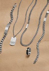 Gioielli in metallo argentato, tra cui un braccialetto strutturato, diverse catene, un ciondolo a forma di targhetta e un anello con un accento nero.