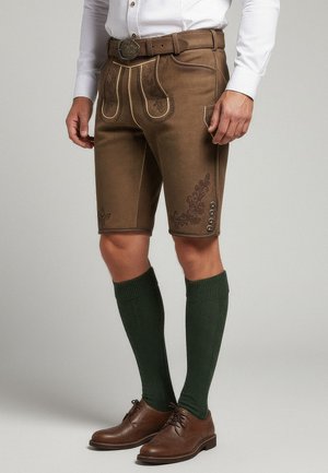 Pantalones cortos de cuero marrón con costuras decorativas, que presentan un ajuste slim. Unos zapatos marrones y unos calcetines verdes hasta la rodilla completan el look.