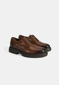 Bruine leren veterschoenen met een ronde neus, voorzien van een zwarte rubberen zool en een minimalistisch ontwerp met stiksels aan de voorkant.