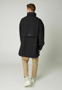 Čierny dlhý kabát s vysokým golierom, s mierne nadmerným strihom, bočnými rozparkami a malým logom hory na chrbte. Béžové nohavice a biele tenisky.