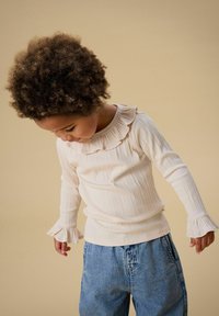 Lys beige ribbestrikket langermet topp med volangkantet utringning og markerte klokkeformede ermer, kombinert med blå denimjeans.