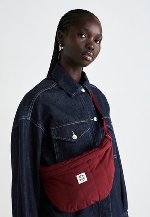 Burgundy quilted shoulder bag with a rectangular shape, featuring a logo patch, worn over a dark denim jacket with two front pockets.