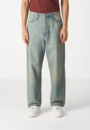 Osoba nosící volné světle modré džíny s vybledlým efektem, bílé tenisky a bordó tričko, stojící před jednobarevným bílým pozadím.