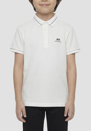 Ragazzo che indossa una polo bianca con rifiniture nere sul colletto e sulle maniche e pantaloni neri, in piedi davanti a uno sfondo semplice.