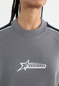 Grå genser med ribbestrikket hals og skulderdetaljer i marineblå. Hvit "COSMO" logo med stjernegrafikk på brystet. Myk tekstur.