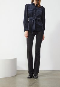 Marineblaue Wildlederbluse mit zwei Brusttaschen, in der Taille mit einem gebundenen Gürtel gerafft. Kombiniert mit schwarzen, ausgestellten Jeans und Stiefeletten.