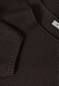 Tissu tricoté marron foncé avec un motif de point texturé visible et une petite étiquette blanche attachée au bord.