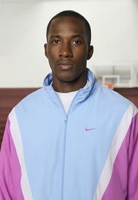 Jonge man die een lichtblauwe en paarse sportjas met ritssluiting draagt, staand binnen met een basketbalring op de vage achtergrond.