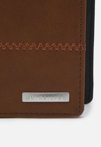 Brun kunstlær lommebok med kontrasterende svart stoff, sømdetaljer og en sølv Quiksilver-logo.