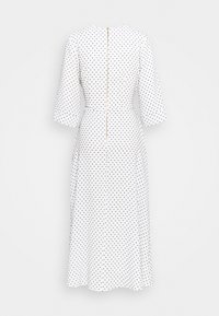 Robe blanche à pois noirs, manches trois quarts, jupe évasée, et fermeture éclair au dos avec une garniture dorée.