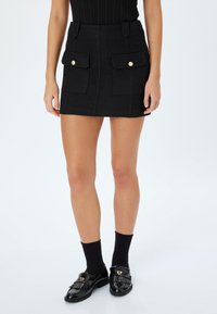 Mini falda negra en tejido texturizado con dos bolsillos frontales, botones dorados y presillas para cinturón, combinada con mocasines negros y calcetines.