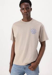 Beige bomull T-skjorte med korte ermer, med et blått grafisk design på venstre brystområde, kombinert med lyseblå jeans.